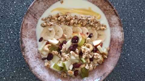 Bild Joghurt Frucht Bowl  Auf diesem Bild befindet sich ein Teller in dessen Mitte Joghurt mit Bananenscheiben, Apfel- und Birnenstücken, Granola und Honig angerichtet ist.