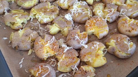 Bild Smashed Potatoes  Auf dem Bild befinden sich mehrere vorgekochte Kartoffeln, die mit einem Glas plattgedrückt und mit geriebenen Käse bestreut sind auf einem Ofenblech