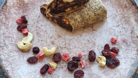 Au diesem Bild befindet sich ein Teller auf dem ein Wrap mit Schokoladenaufstrich und Bananen gefüllt ist. Der Teller ist mit Nüssen und Obst verziert.